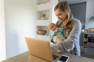 Cheerful new mom rocking baby and showing content on laptop to her. Portrait of young woman and cute little child in home interior. Wireless connection concept